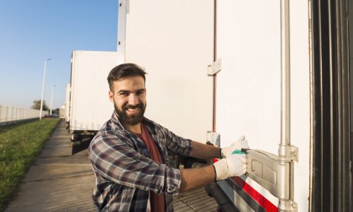 Smiling truck driver in working gloves opening or closing truck trailer back doors checking goods for transportation.